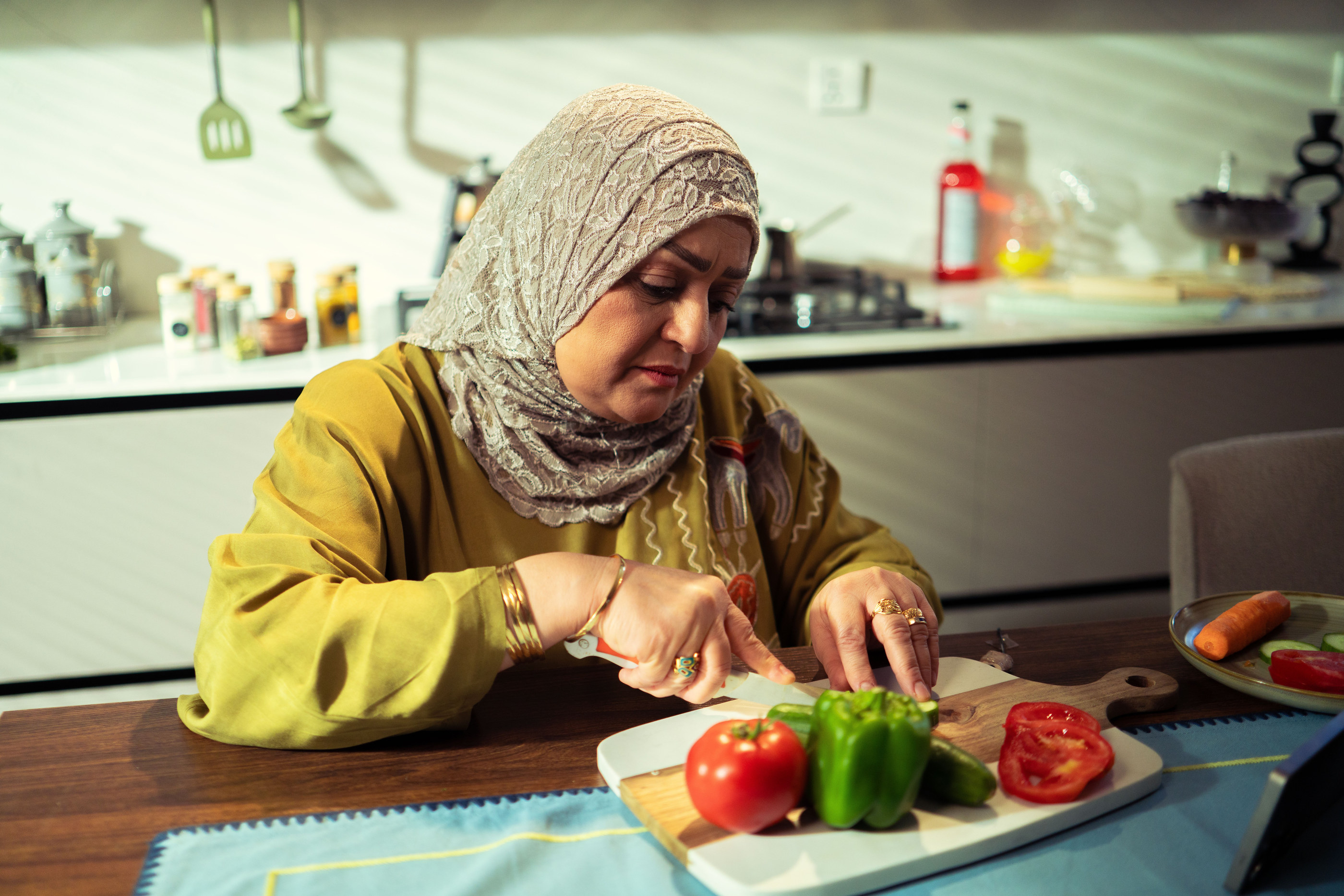 A woman in a headscarf chopping peppers and tomatoes in her kitchen.