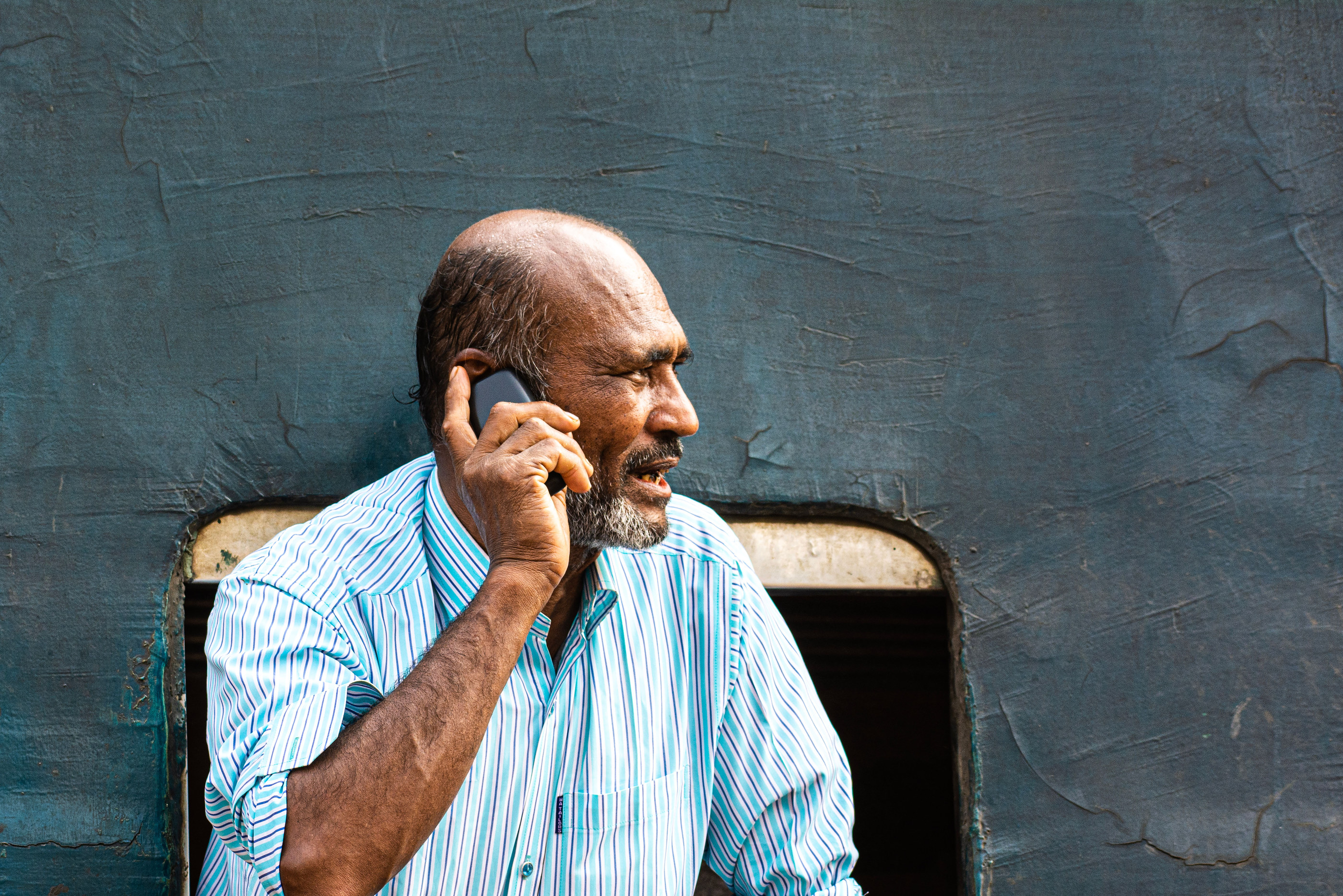 A man in his sixties taking a call at home, seated by a window.